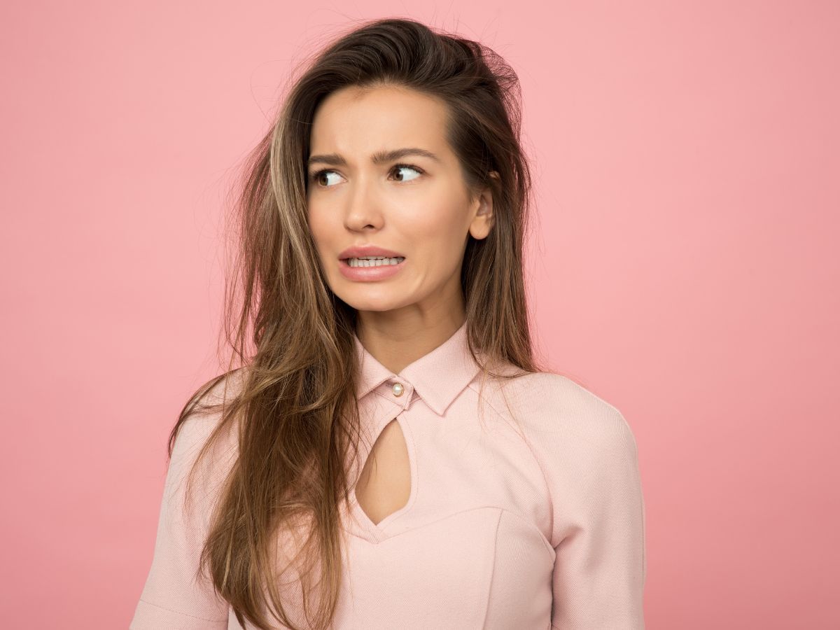 An image of a woman dressed in a light pink top in front of a pink background looking unsure and nervous.